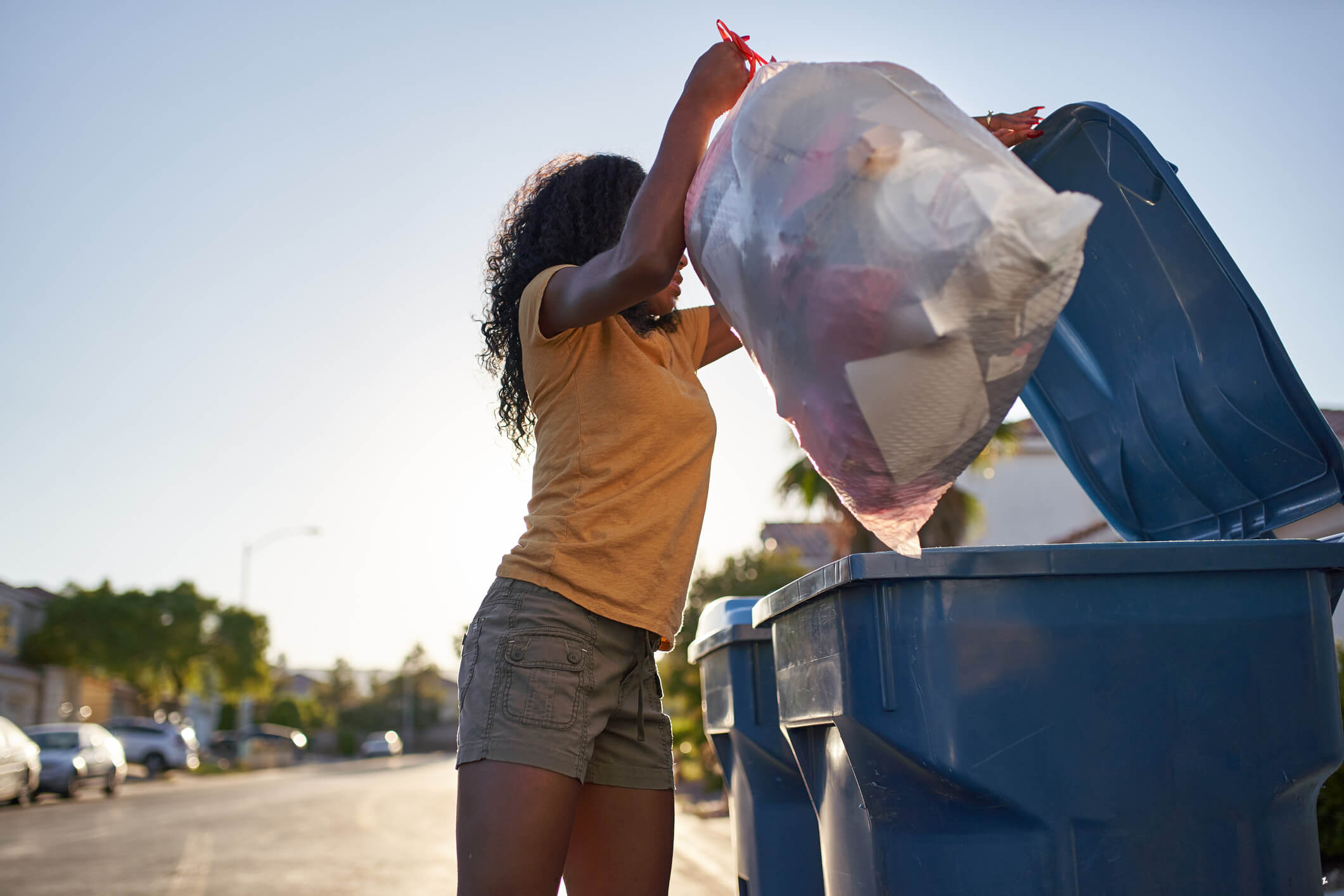 Shows a lady placing a full bag of rubbish into a wheelie bin